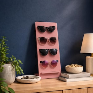 Puede incluir: Un expositor vertical de madera en color rosa empolvado con cuatro pares de gafas de sol dispuestas en columna. El soporte cuenta con una base rectangular plana y un pequeño borde horizontal en la parte inferior para sujetar las gafas de forma segura.