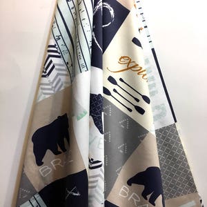 May include: Fabric with a patchwork design featuring bears, arrows, and geometric patterns in navy, tan, gray, and white. The word "BRAVE" is printed near a bear silhouette. The word "Explore" is printed in a gold color. The fabric is draped.