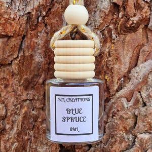 May include: A small glass bottle with a wooden top and a hanging string. The label reads "BCL CREATIONS BLUE SPRUCE 8ML". The bottle is suspended against a textured brown background.