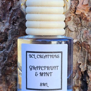 May include: A clear glass bottle with a wooden cap and a white label that reads "BCL Creations Grapefruit & Mint 8ML". The bottle is hanging from a gold and white braided string.
