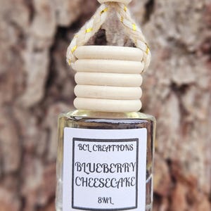 May include: A small, clear glass bottle with a wooden cap and a white label. The label reads "BCL Creations Blueberry Cheesecake 8mL".