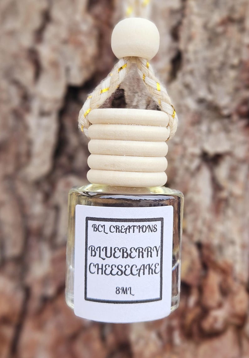 May include: A small, clear glass bottle with a wooden cap and a white label. The label reads "BCL Creations Blueberry Cheesecake 8mL".