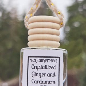 May include: A clear glass bottle with a white label that reads "BCL Creations Crystallized Ginger and Cardamom 8ML". The bottle is hanging from a wooden bead and twine.