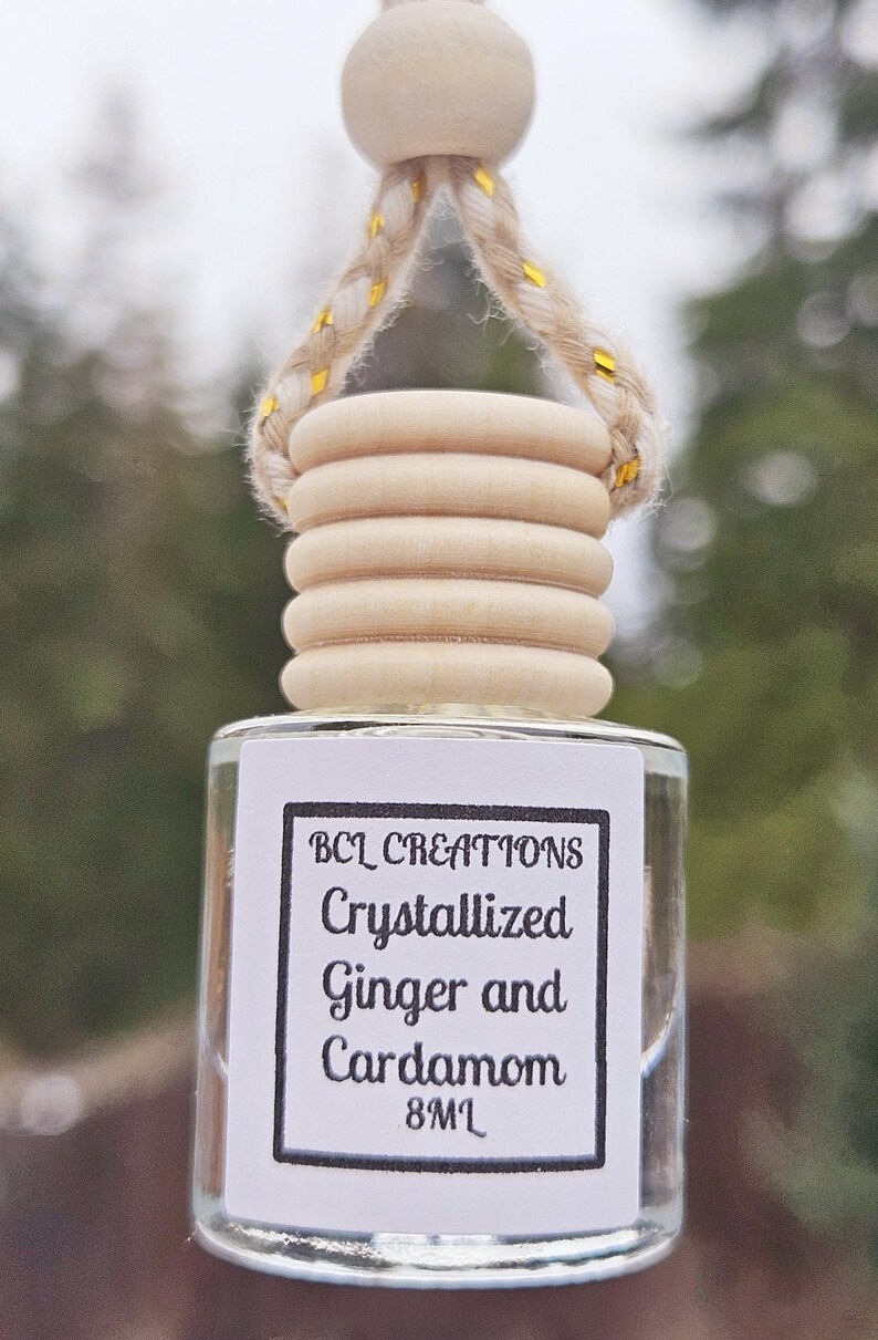 May include: A clear glass bottle with a white label that reads "BCL Creations Crystallized Ginger and Cardamom 8ML". The bottle is hanging from a wooden bead and twine.