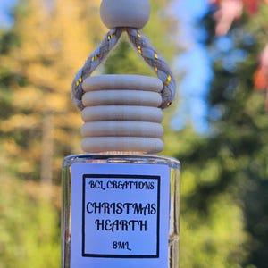 May include: A clear glass bottle with a white label that reads "BCL Creations Christmas Hearth 8ml". The bottle is hanging from a white rope with gold accents.
