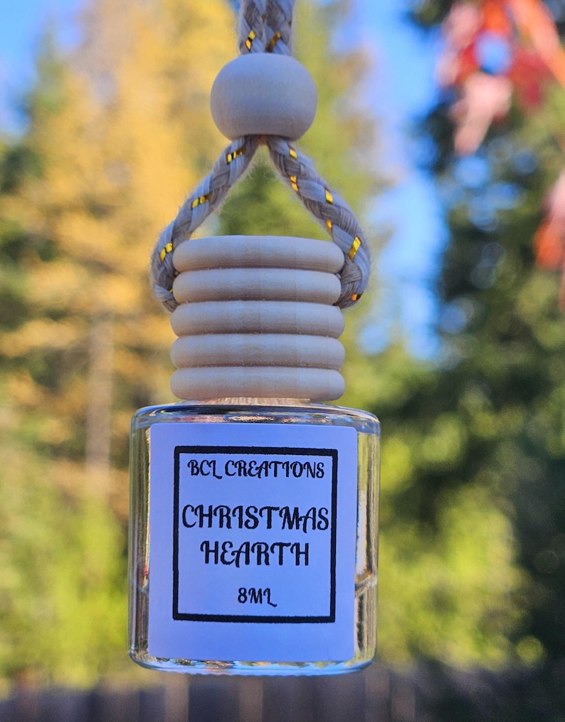May include: A clear glass bottle with a white label that reads "BCL Creations Christmas Hearth 8ml". The bottle is hanging from a white rope with gold accents.