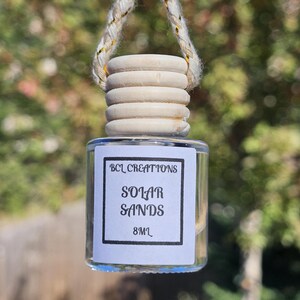 May include: A clear glass bottle with a wooden cap and a white label that reads "BCL Creations Solar Sands 8ml". The bottle is hanging from a string.