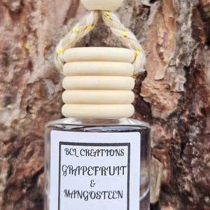 May include: A clear glass bottle with a wooden cap and a white label that reads "BCL Creations Grapefruit & Mangosteen 8ml". The bottle is hanging from a string with a wooden bead.