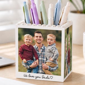May include: A white square pen holder with a top rim resembling building blocks. The front features a photograph of a man holding two young children, with the text "We love you Dada" and a heart symbol printed in black at the bottom.