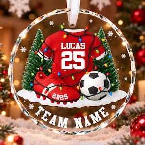 May include: A round, clear acrylic ornament featuring a red soccer jersey with the name LUCAS and number 25, a red soccer cleat with 2025, and a soccer ball. The design includes green pine trees, white snow, and string lights, with the text TEAM NAME.