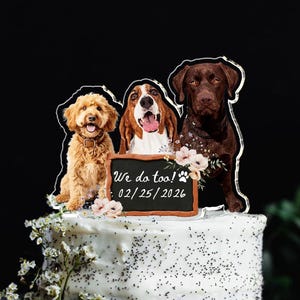 May include: A white frosted cake topped with a clear acrylic topper featuring three dogs and a small chalkboard sign that reads We do too! 02/25/2026. The cake is decorated with small white flowers and green leaves at the base of the topper.