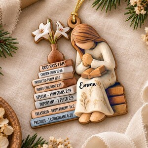 May include: A wooden hanging ornament featuring a figure with brown hair in a cream-colored dress holding a book, sitting on a stack of books labeled with Bible verses and the name Emma, topped with a vase containing two white lily flowers.