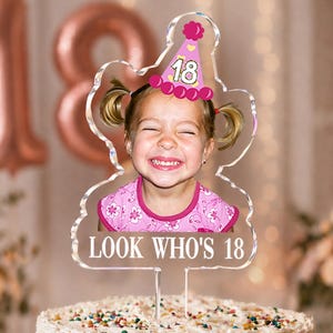 May include: A clear acrylic cake topper featuring a photograph of a smiling child wearing a pink party hat with the number 18. The topper includes the white text LOOK WHO'S 18 printed at the bottom and is positioned on a frosted cake with sprinkles.