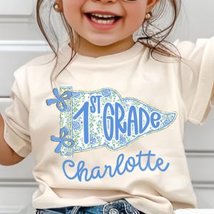 May include: A cream-colored t-shirt featuring a blue and white floral design with the text "1st Grade" and the name "Charlotte" in blue. The shirt has short sleeves and is made of a soft material.