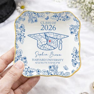 May include: A white ceramic trinket dish with a blue and gold floral design. The dish features the text "CLASS OF 2026", a graduation cap map, the name "Sophia Brown", and "HARVARD UNIVERSITY" with coordinates. The dish is held by a hand.