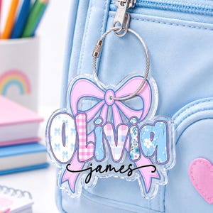 May include: A clear acrylic keychain featuring a large pink bow graphic behind the name Olivia in blue and pink patterned letters, with the name james written in black cursive script below, attached to a silver metal cable ring on a light blue bag.