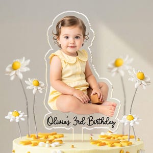May include: A cake topper featuring a photo of a baby girl in a yellow outfit, with the text "Olivia's 3rd Birthday." The cake is decorated with daisies and sun-shaped accents, creating a cheerful, celebratory theme.