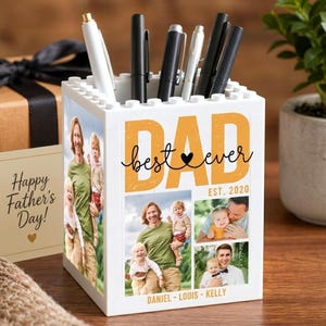May include: White block-style pen holder with the words "DAD best ever" in orange and black. The pen holder has photos of a family. The words "Happy Father's Day!" are on a card next to the pen holder.