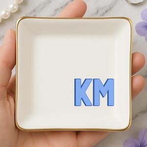 May include: A square, off-white ceramic trinket dish with a gold rim, featuring the blue letters "KM". The dish is held in a hand, with a string of pearls and a purple flower in the background. A decorative item for jewelry or small items.