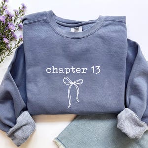 May include: A blue-grey sweatshirt with the text "chapter 13" and a bow graphic in white. The sweatshirt has a crew neck and ribbed cuffs. A sprig of purple flowers is visible in the upper left corner.