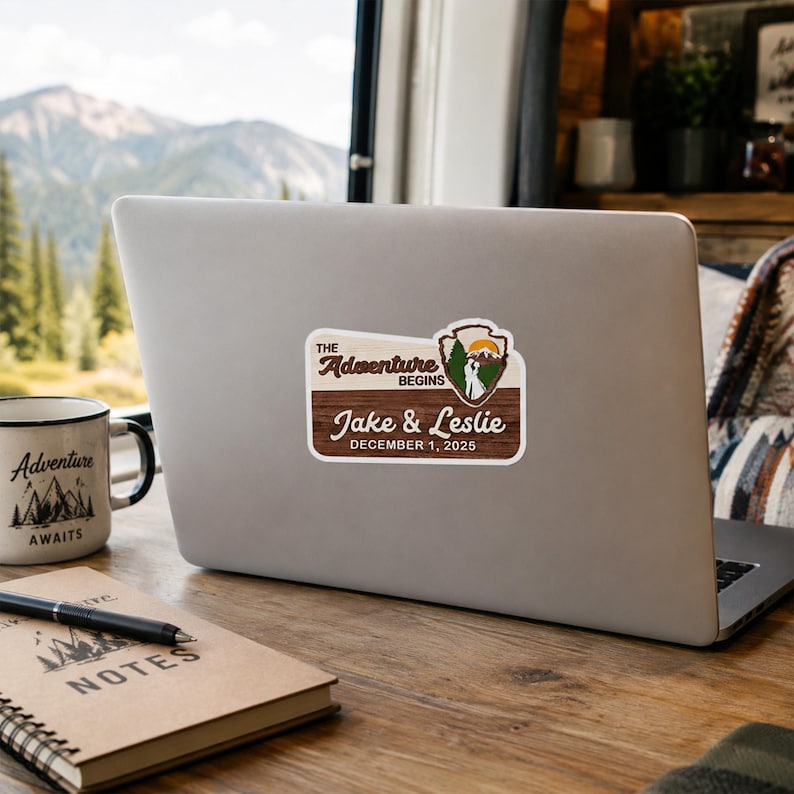 Puede incluir: Una pegatina rectangular con textura de veta de madera que presenta el texto "THE ADVENTURE BEGINS", "Jake & Leslie" y "DECEMBER 1, 2025". Incluye un gr&aacute;fico de un paisaje monta&ntilde;oso dentro de un emblema en forma de escudo.
