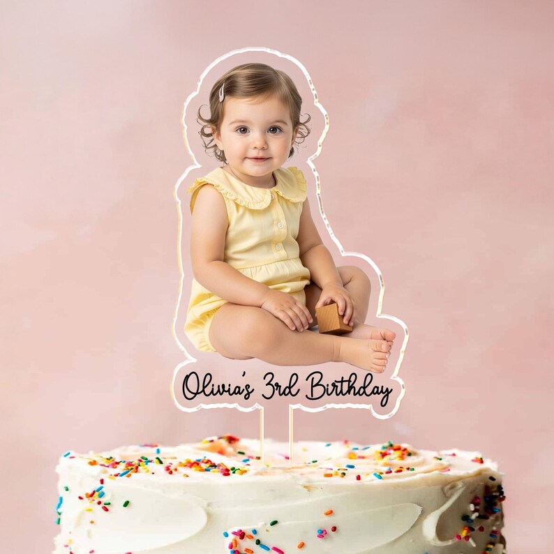 May include: A cake topper featuring a photo of a young child in a yellow outfit, with the text "Olivia's 3rd Birthday." The topper is placed on a white frosted cake decorated with colorful sprinkles. The background is a soft pink.