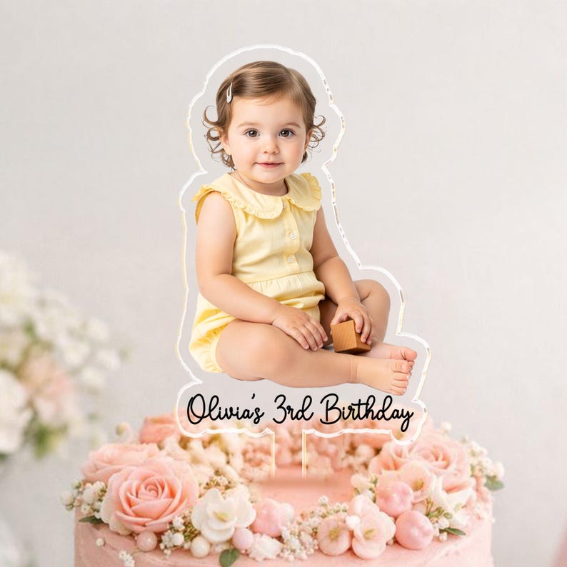 May include: A cake topper featuring a photo of a young child in a yellow outfit, with the text "Olivia's 3rd Birthday." The topper is placed on a pink cake decorated with pink flowers and greenery.