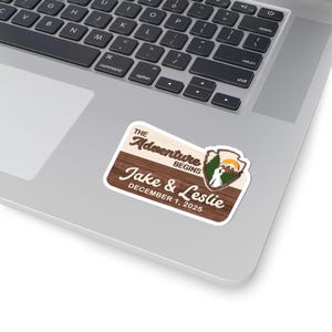 Puede incluir: Una pegatina rectangular con fondo de veta de madera que presenta el texto The Adventure Begins, Jake & Leslie, y December 1, 2025. Un peque&ntilde;o emblema a la derecha muestra un paisaje de monta&ntilde;a con un sol, &aacute;rboles y dos siluetas humanas.