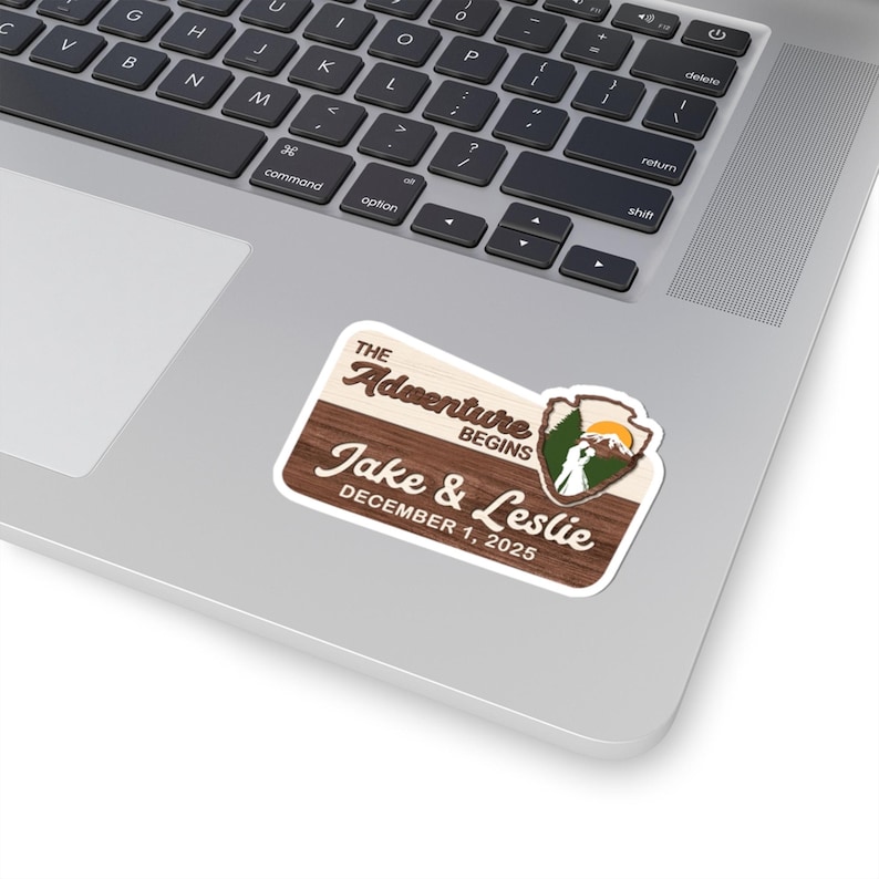 Puede incluir: Una pegatina rectangular con fondo de veta de madera que presenta el texto The Adventure Begins, Jake & Leslie, y December 1, 2025. Un peque&ntilde;o emblema a la derecha muestra un paisaje de monta&ntilde;a con un sol, &aacute;rboles y dos siluetas humanas.
