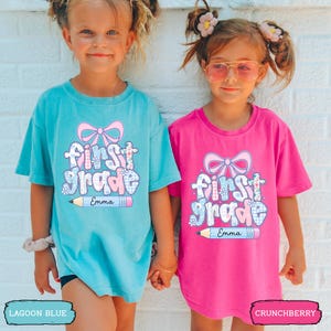 May include: A light pink t-shirt featuring a graphic print with a large bow, the text "first grade" in a blue and white plaid pattern, and a pencil illustration at the bottom with the name "Emma" written in black script on the pencil shaft.