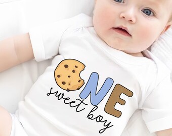 May include: A white onesie with a blue and brown "ONE" graphic and a chocolate chip cookie with a bite taken out of it. The text "sweet boy" is written below the number.