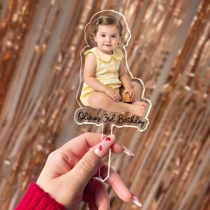 May include: Clear acrylic cake topper featuring a photo of a young child in a yellow outfit. The text "Olivia's 3rd Birthday" is written in a cursive font. The topper is held by a hand with red nail polish.