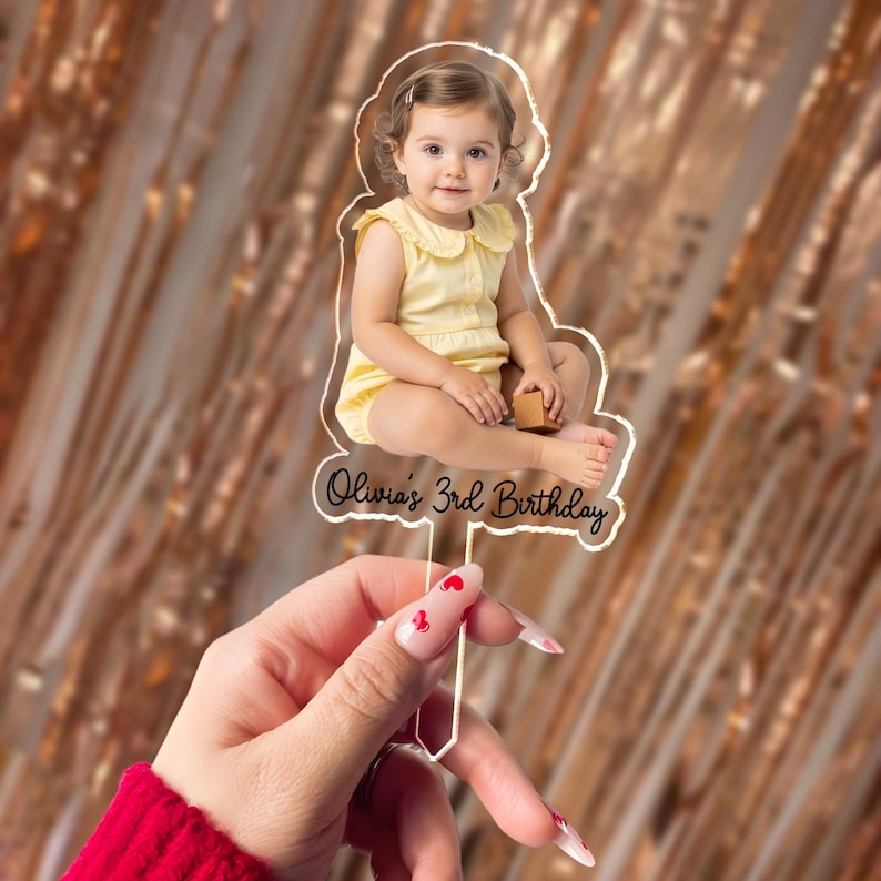 May include: Clear acrylic cake topper featuring a photo of a young child in a yellow outfit. The text "Olivia's 3rd Birthday" is written in a cursive font. The topper is held by a hand with red nail polish.