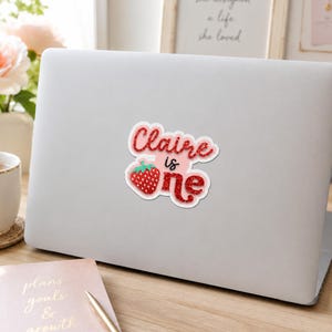 Puede incluir: Una pegatina troquelada con el texto Claire is one en acabado de purpurina roja. La palabra Claire y la palabra one est&aacute;n escritas en fuente cursiva, mientras que is est&aacute; en letra negra simple. Un gr&aacute;fico de fresa sustituye a la letra o.