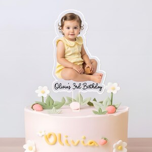 May include: A light pink birthday cake decorated with white flowers, green leaves, and strawberry accents. A cake topper features a photo of a child and the text "Olivia's 3rd Birthday".