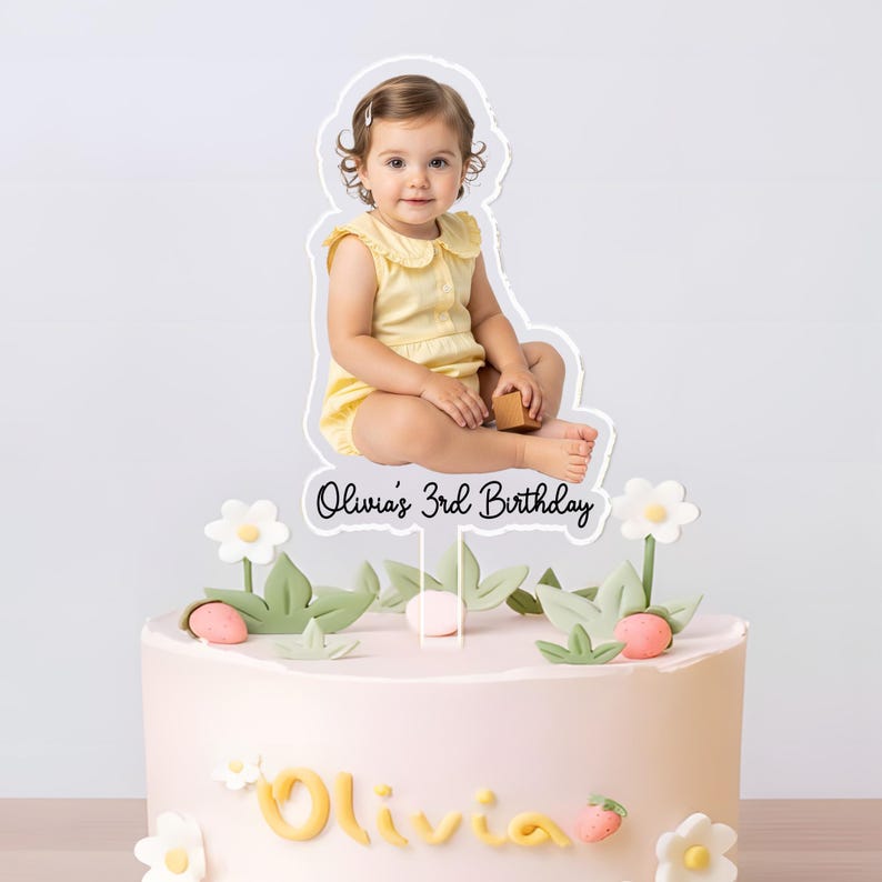 May include: A light pink birthday cake decorated with white flowers, green leaves, and strawberry accents. A cake topper features a photo of a child and the text "Olivia's 3rd Birthday".