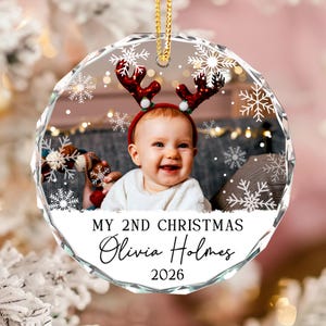 May include: A round, faceted glass Christmas ornament featuring a photo of a baby wearing red reindeer antlers. The ornament is decorated with white snowflakes and includes the text: MY 2ND CHRISTMAS, Olivia Holmes, and the year 2026.