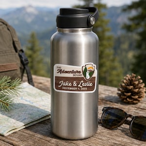 Puede incluir: Una botella de agua de acero inoxidable con tapa de rosca negra y una pegatina personalizada. La pegatina muestra un gr&aacute;fico de monta&ntilde;a, el texto "THE Adventure BEGINS", "Jake & Leslie" y la fecha "DECEMBER 1, 2025" sobre un fondo de madera.