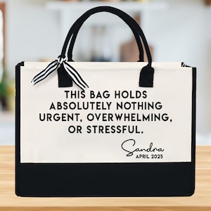 May include: A canvas tote bag with black handles and a black bottom panel. The bag has the text "THIS BAG HOLDS ABSOLUTELY NOTHING URGENT, OVERWHELMING, OR STRESSFUL." with the name "Sandra" and the date "APRIL 2025" printed on it.
