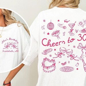 May include: White t-shirt with pink text and illustrations. The front of the shirt says "Alex's Birthday Scottsdale, AZ". The back of the shirt says "Cheers to 30!" with illustrations of wine glasses, a pizza, a heart-shaped cake, and fireworks.