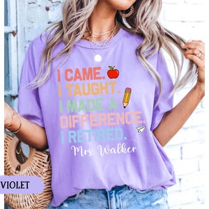 May include: Purple t-shirt with the text "I came. I taught. I made a difference. I retired. Mrs. Walker" in rainbow colors with a red apple, pencil, and paper airplane graphic.