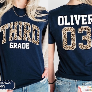 May include: Navy blue t-shirt with "THIRD GRADE" in a leopard print design on the front. The back of the shirt displays "OLIVER" and the number "03" in a leopard print design. The shirt is a Bella Canvas brand.