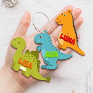 May include: Three colorful dinosaur-shaped keychains. One is green with the name "LISA", another is blue with "AVA", and the third is orange with "LUNA". Each keychain has a silver metal loop for attachment. These are likely personalized accessories.