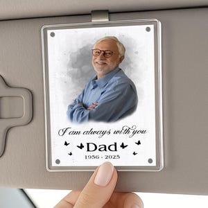 May include: A rectangular, clear acrylic car visor ornament featuring a photo of a man wearing glasses and a blue shirt. The text reads "I am always with you Dad 1956-2025" with butterfly accents. The ornament is held by a hand.