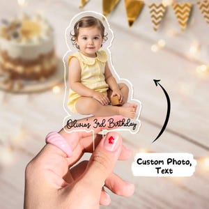 May include: A personalized cake topper featuring a photo of a young child in a yellow outfit. The text "Olivia's 3rd Birthday" is displayed below the image. The topper is held by a hand with a pink ring and a red heart nail design.