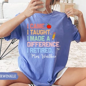 May include: A light blue t-shirt with the text "I came, I taught, I made a difference, I retired. Mrs. Walker" in rainbow colors. The text is surrounded by a red apple, a yellow pencil, and a paper airplane.