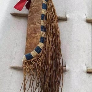 Könnte beinhalten: Eine Lederhülle im indianischen Stil mit einem blau-beigen Perlendesign. Die Hülle hat lange, braune Fransen und einen roten Stoffakzent. Sie ist an einem Holzdübel aufgehängt.