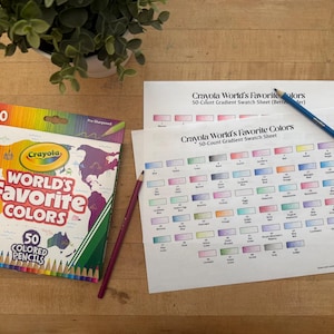 May include: A box of Crayola World's Favorite Colors 50 colored pencils with a world map design. Two pencils and two color gradient swatch sheets are also visible. A potted plant sits in the background on a wooden surface.