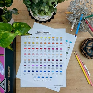 Może przedstawiać: Arkusz wzorów kolorów Prismacolor z 150 kolorami w kształcie owali. Arkusz leży na drewnianej powierzchni z rośliną w doniczce, ołówkami i drewnianym pudełkiem.