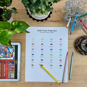 Op de afbeelding: Een wit vel papier met een kleurenkaart voor Koh-I-Noor Polycolor 24 kleuren potloden. De kaart toont 24 verschillende kleurstalen met de bijbehorende kleurennamen en nummers. Er liggen ook drie kleurpotloden op de tafel.